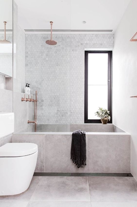 Banheiro com banheira de alvenaria revestida de porcelanato, revestimento marmorizado hexagonal e metais bronze.